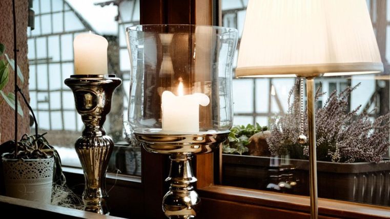 Ein stilvolles Arrangement mit Kerzen und einer Lampe auf einem Tisch. Im Hintergrund sind Fenster und Pflanzen sichtbar.