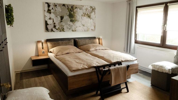 Ein modernes Schlafzimmer mit einem Doppelbett und hellen Bettdecken. Die Wände sind dezent gestaltet, und große Fenster sorgen für natürliche Beleuchtung.
