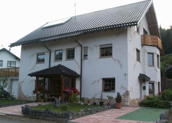 A large, two-story house with a black roof and an ornate facade. In the foreground, there is a garden and plant containers.