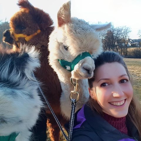 Eine Frau lächelt mit zwei Alpakas im Hintergrund. Die Alpakas tragen Halsbänder und scheinen freundlich und neugierig zu sein.
