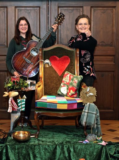 Two people are standing next to a decorated chair. One of them is holding a guitar, and both are smiling happily.