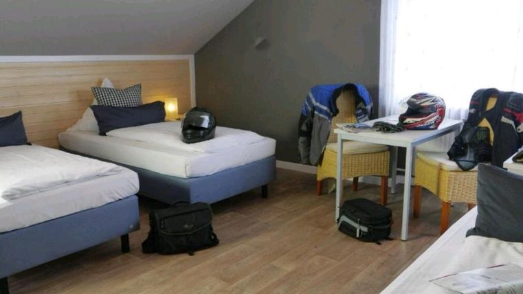 A simple room with two single beds and a table. Motorcycle gear and helmets are scattered throughout the room.
