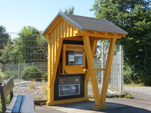 Een geel verkooppunt met een schuin dak staat op een perron. Naast het verkooppunt is er een bank en een hekje op de achtergrond.