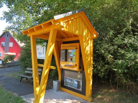 Eine gelbe Verkaufsstelle mit einem Vordach steht in einer ruhigen Umgebung. Im Hintergrund sind Bäume und ein rotes Gebäude sichtbar.