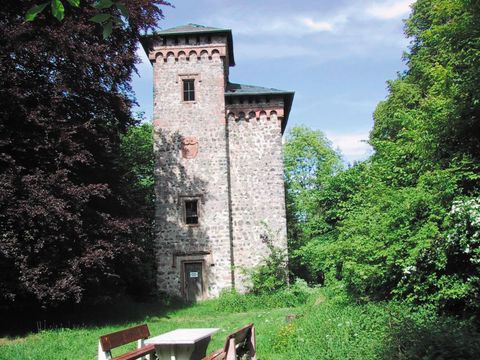 Ein alter Turm aus Stein, umgeben von üppigem Grün und Bäumen. Im Vordergrund stehen eine Bank und ein Tisch in der Natur.