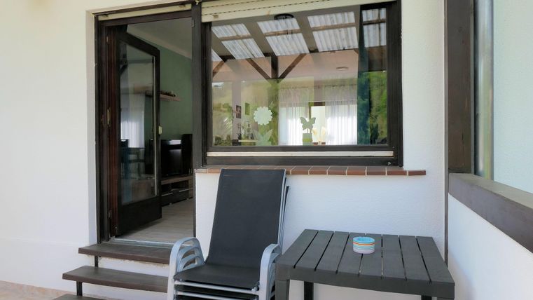 Eine gemütliche Terrasse mit einem Liegestuhl und einem kleinen Tisch. Der Blick geht durch ein großes Fenster ins Innere des Raumes.