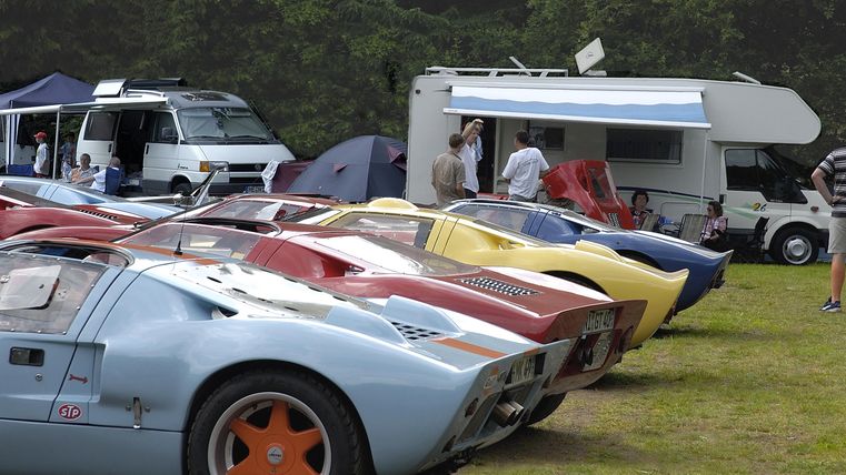 A collection of classic sports cars is parked on a meadow. In the background, there are motorhomes and people visible.