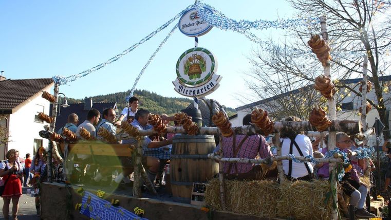 Ein festlicher Umzugswagen mit fröhlichen Menschen in traditioneller Kleidung. Es sind Bierfässer, Brezeln und Dekorationen zu sehen, die eine festliche Atmosphäre schaffen.