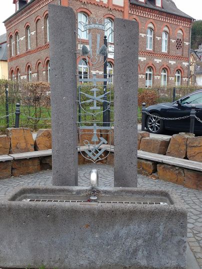 Ein moderner Brunnen mit zwei vertikalen Steinsäulen steht in einem kleinen Platz. Im Hintergrund ist ein historisches rotes Gebäude zu sehen.