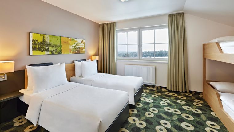 A modern hotel room with two single beds and a bunk bed. The room is bright and friendly, with a large window and a stylish carpet.