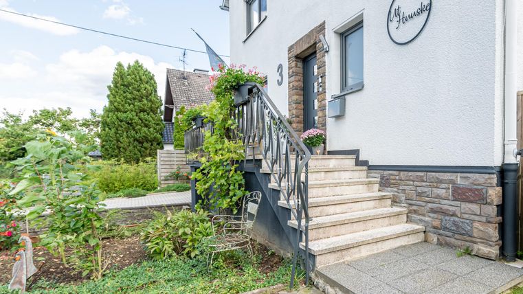 Een modern huis met een uitnodigende trap en groene beplanting. De lucht is helder en blauw, wat een vriendelijke sfeer creëert.