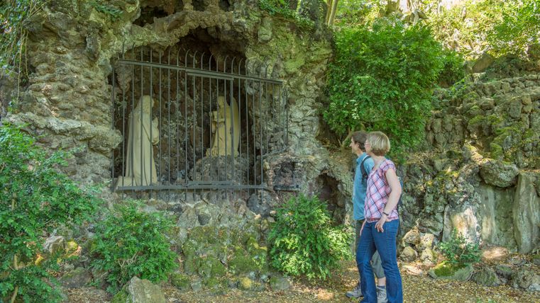 Zwei Personen betrachten eine in Felsen eingelassene Grotte mit Statuen, die durch ein Gitter geschützt sind.