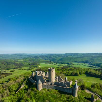 Eine beeindruckende Burgruine auf einem hügeligen Gelände. Rundherum erstrecken sich grüne Wiesen und ein klarer blauer Himmel.