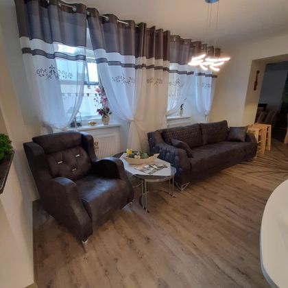 A modern living room with a comfortable sofa and an armchair. Large windows with light curtains provide plenty of daylight.