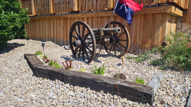 Een historisch kanon staat in een stenen tuin, omringd door planten. Op de achtergrond is een houten hek en een vlag te zien.