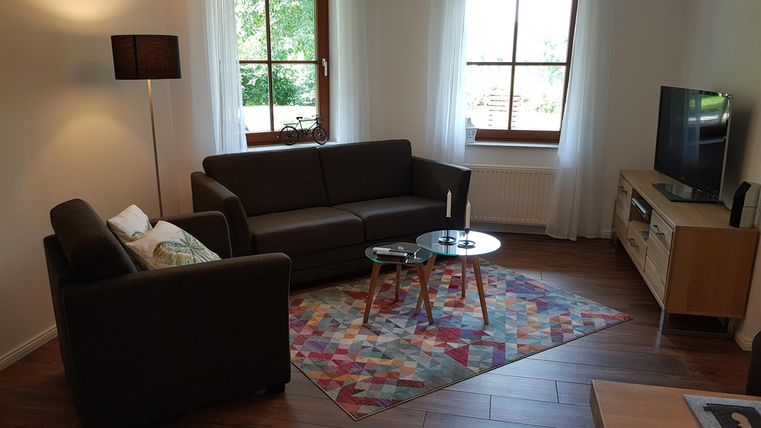 Ein helles Wohnzimmer mit einer dunklen Couch und einem Sessel. Ein runder Tisch und ein Teppich mit buntem Muster ergänzen die gemütliche Atmosphäre.