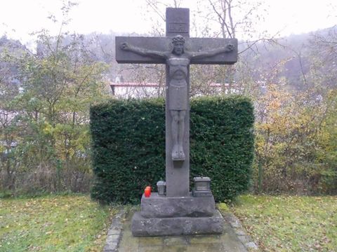 Ein Kreuz mit einer Christusfigur steht in einer grünen Umgebung. Der Himmel ist bewölkt und die Landschaft wirkt ruhig.