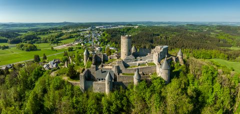 Weitblick über die Burgruine Nürburg mit ihren massiven Steinmauern und Türmen, umgeben von dichtem Grün und einer weitläufigen Hügellandschaft. Im Hintergrund erstrecken sich Felder, Wälder und ein kleiner Ort unter klarem Himmel.