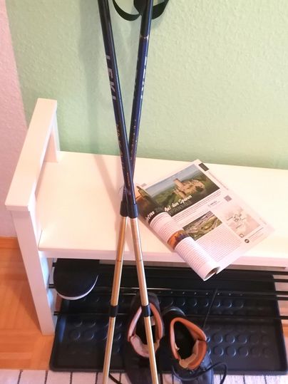 Zwei Wanderstöcke stehen auf einem Sitzbank. Neben ihnen liegen Wanderschuhe und eine Zeitschrift.