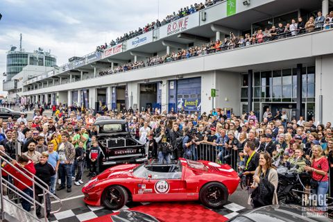 Ein großer Menschenauflauf bei einer Automobilveranstaltung. Im Vordergrund steht ein rotes Rennfahrzeug, während sich zahlreiche Zuschauer auf den Tribünen versammeln.