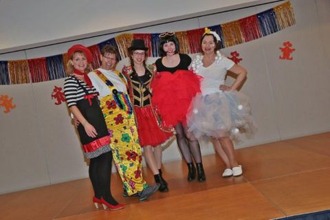 Vijf vrouwen staan in kleurrijke kostuums op een podium. Op de achtergrond zijn feestelijke versieringen te zien.