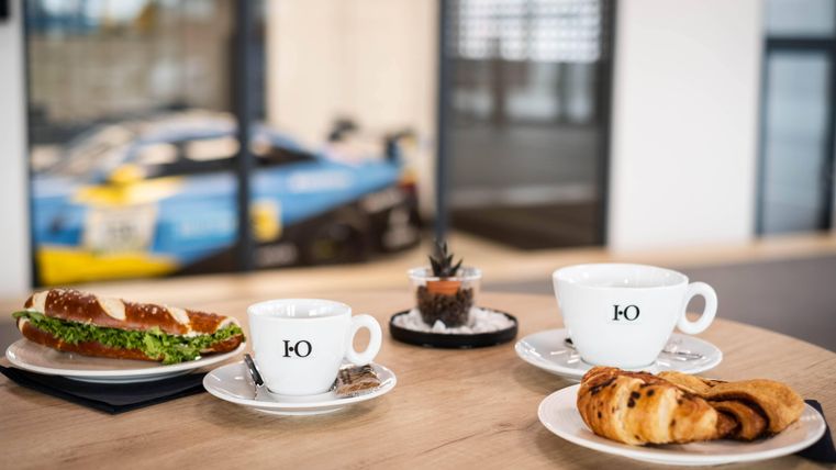 Ein Tisch mit zwei Kaffeetassen, einer Brezel und einem Croissant. Im Hintergrund ist ein Rennwagen sichtbar.
