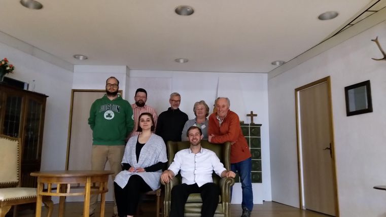 Eine Gruppe von sieben Personen steht und sitzt in einem Zimmer. Es gibt Möbel und eine helle, freundliche Atmosphäre.