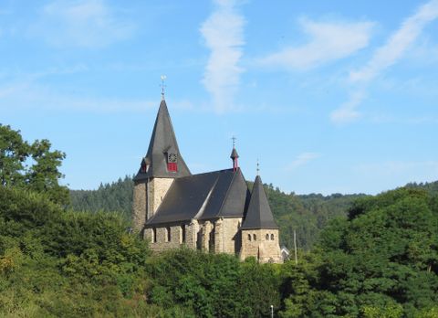 Une vieille église entourée d'arbres et de collines. Le ciel est bleu avec quelques nuages.