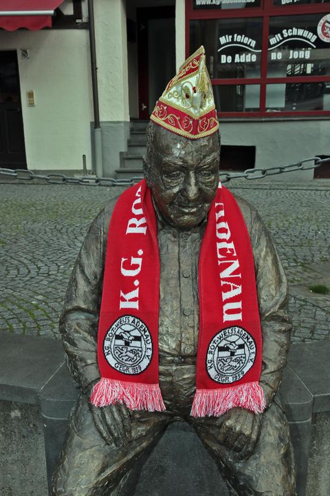 Eine Bronzestatue eines sitzenden Mannes, der einen roten Schal und eine festliche Mütze trägt. Die Statue ist auf einem Pflastersteinplatz in der Stadt aufgestellt.