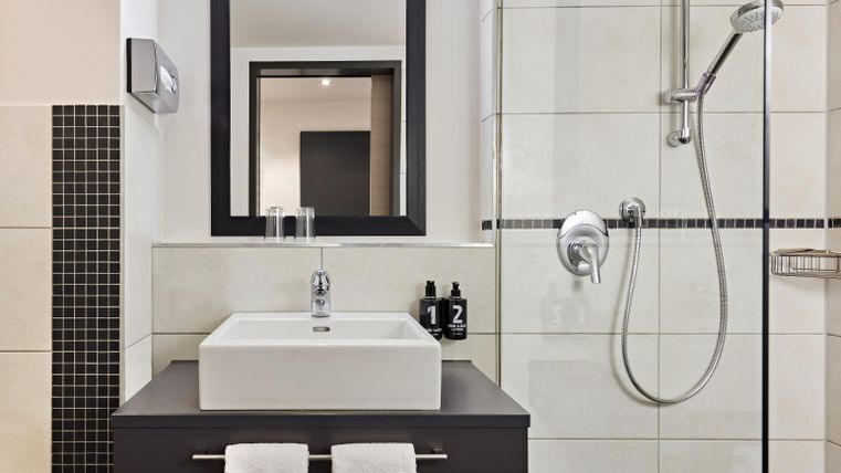 A modern bathroom with a rectangular sink and a large mirror. The shower is separated by a glass wall, and two towels are ready.