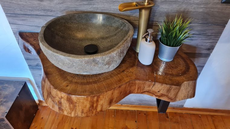 Een unieke wastafel van natuurlijk hout met een ronde stenen kom. Daarop staan een gouden kraan, een zeepdispenser en een kleine pot met groenplanten.