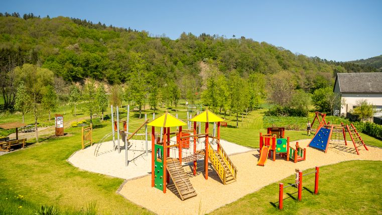 Spielplatz in Müsch mit bunten Klettergerüsten und Rutschen, umgeben von grüner Landschaft und Bäumen.