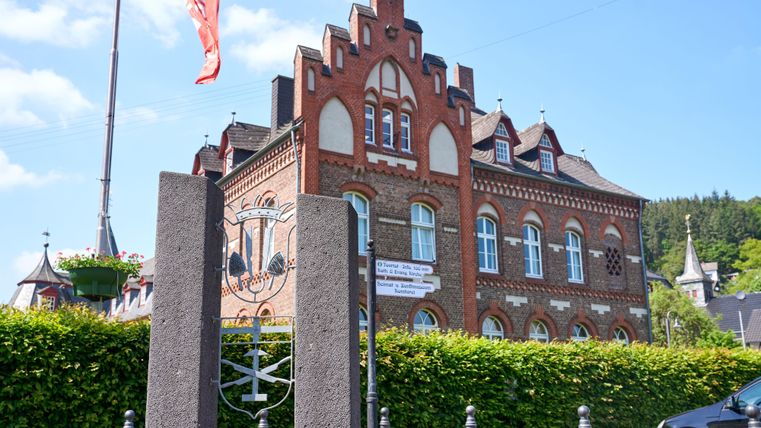 A historic building with a striking red facade and beautiful windows. In the foreground, a fence and a pathway can be seen, surrounded by green shrubs.
