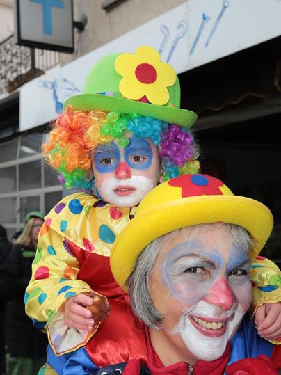 Ein fröhlicher Clown trägt ein Kind auf den Schultern. Beide haben bunte Kostüme und geschminkte Gesichter.