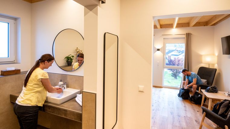 A modern bathroom with a sink and a large mirror. In the background, a cozy living area is visible where a person is relaxing.