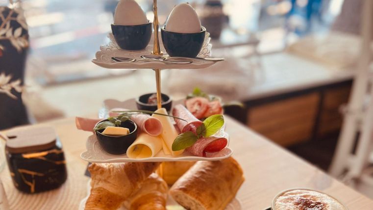 Un arrangement de petit déjeuner élégant avec deux œufs, de la charcuterie, du fromage, des herbes fraîches et des pâtisseries sur un présentoir moderne. Accompagné d'une tasse de café avec de la mousse.