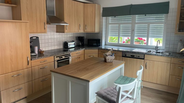 Een moderne keuken met houten kasten en een grote tafel in het midden. Heldere ramen laten veel licht binnen, en er is uitzicht op bloeiende planten buiten.