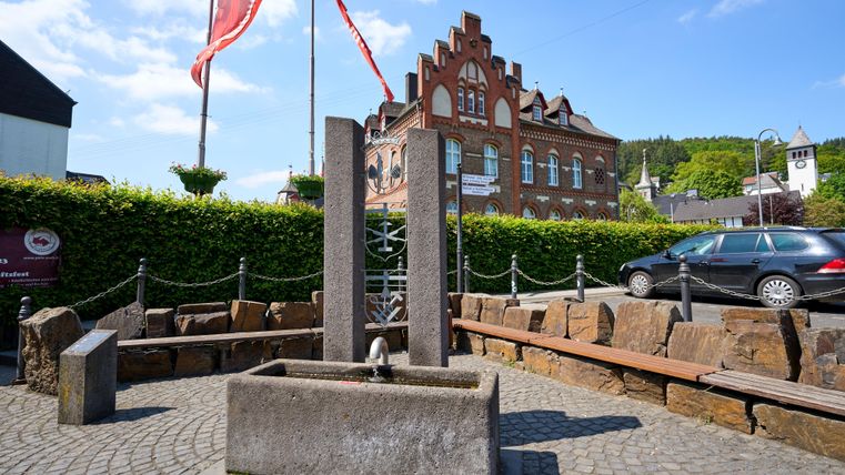 Een mooie plek met een stenen fontein en natuurstenen banken. Op de achtergrond is een historisch gebouw en vlaggen te zien.