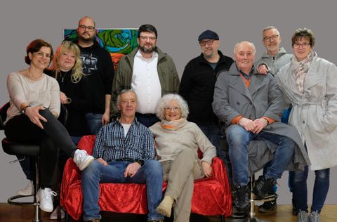 Eine Gruppe von zehn Personen sitzt und steht vor einem neutralen Hintergrund. Sie lächeln und wirken freundlich in einer entspannten Atmosphäre.