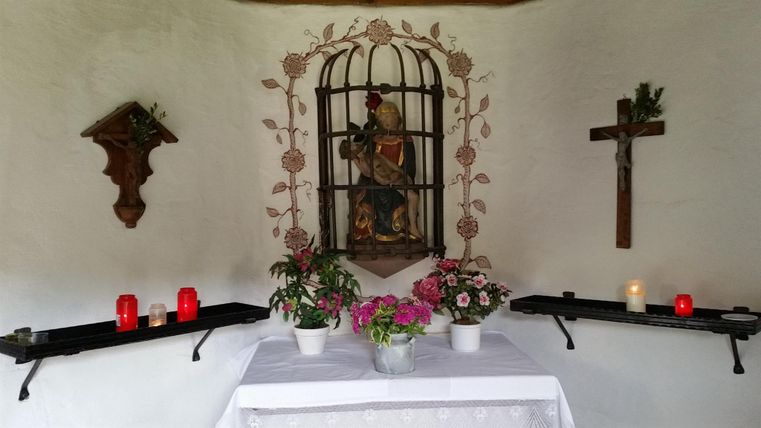 Eine kleine Kapelle mit einer Statue in einem Gitter und Blumenarrangements auf einem Tisch. An den Wänden sind Kerzen und ein Kreuz zu sehen.