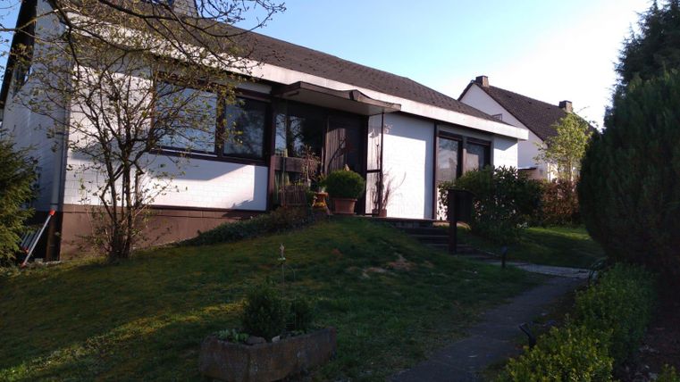 A simple house with a well-maintained garden and trees. The sky is clear and it is a sunny day.