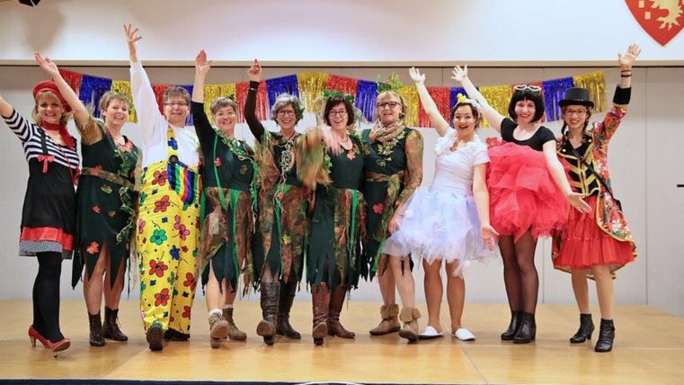 Een groep vrouwen in kleurrijke kostuums staat op een podium en steekt de armen in de lucht. Ze zien er vrolijk uit en vieren een feestelijk evenement.