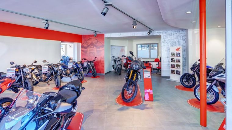 Ein moderner Motorradladen mit verschiedenen Motorrädern in unterschiedlichen Farben. Die Wände und der Boden sind überwiegend in neutralen Tönen gehalten, mit roten Akzenten.