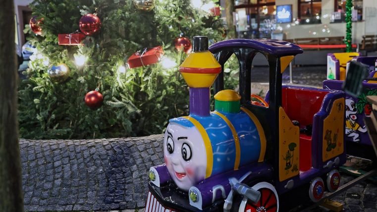 Eine bunte Miniatur-Eisenbahn steht vor einem festlich geschmückten Weihnachtsbaum. Im Hintergrund sind Lichter und Weihnachtsdekorationen zu sehen.
