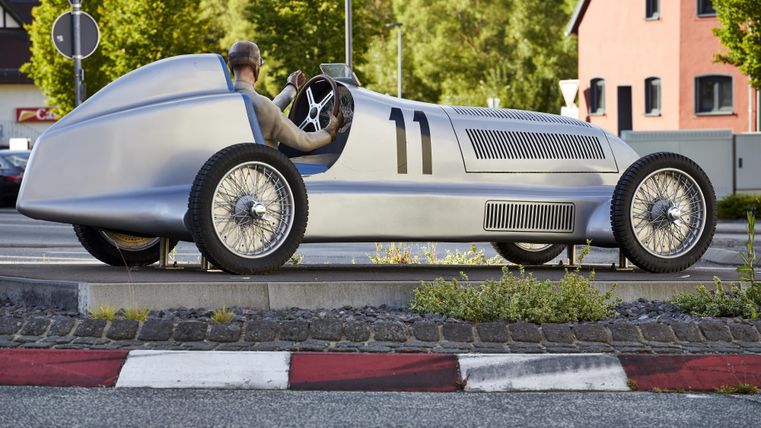 A historic race car with the license plate 11 stands on a road. The driver is sitting in the car and it is surrounded by trees and residential houses.