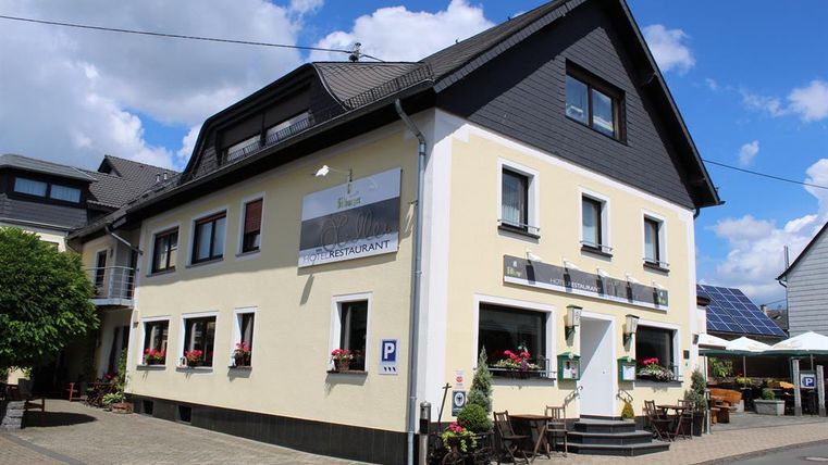 A modern restaurant building with blooming windows and a parking lot in front. The facade is bright and welcoming, surrounded by a beautiful landscape.
