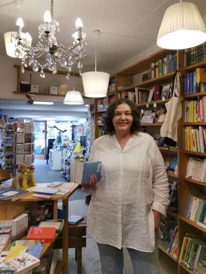 A friendly woman stands in a cozy bookstore. Around her are many bookshelves and a beautiful ceiling light.