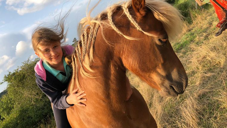 Une fille est assise sur un cheval brun et sourit. À l'arrière-plan, on voit des collines et un environnement rural.