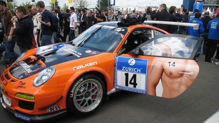 Une voiture de course orange frappante avec des inscriptions publicitaires est garée sur la piste de course. Une foule est rassemblée autour d'elle.