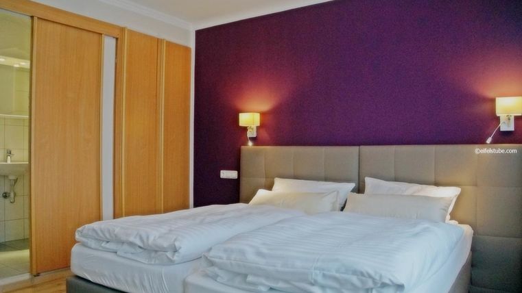 Une chambre moderne avec deux lits simples et un mur violet. À l'arrière-plan, un placard fermé et une salle de bain sont visibles.